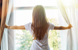 A woman stands in front of a window, arms wide open, inviting the fresh air and sunlight into her space.