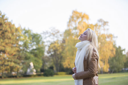 Junge blonde Frau in schöner Herbstnatur atmet tief durch und hat dabei die Augen geschlossen. Die Sonne scheint, sie steht auf einer Wiese und die Blätter der Bäume sind leicht rot gefärbt.