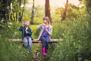 Zwei Kinder auf einer Bank im Wald, die fröhlich Seifenblasen pusten, umgeben von grünen Bäumen und der Natur.