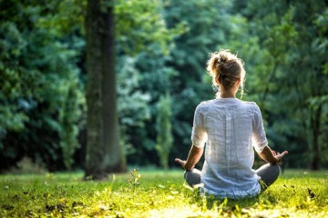 Eine Frau meditiert im Gras vor Bäumen und genießt die Ruhe der Natur in einer friedlichen Umgebung.