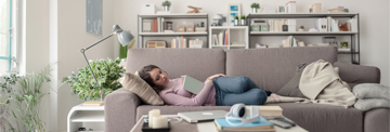 Eine Frau sitzt auf einem Sofa vor einem Bücherregal und liest entspannt ein Buch.