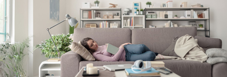 Eine Frau sitzt auf einem Sofa vor einem Bücherregal und liest entspannt ein Buch.