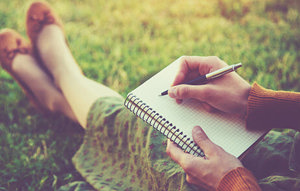 A woman is engaged in writing in her notebook while seated on the grass, enjoying a peaceful outdoor setting.