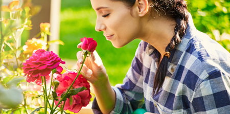 Eine Frau in einem blau-weiß karrierten Hemd, die an einer Rose eines Rosenbuschs riecht.